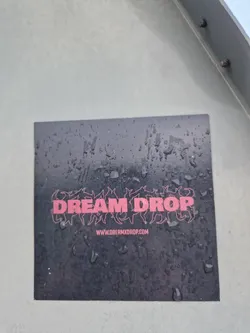 A black sticker with the words DREAM DROP in pink stylized text. Below is a website address: www.loremxdrop.com. The sticker appears to be wet, with water droplets visible on its surface.