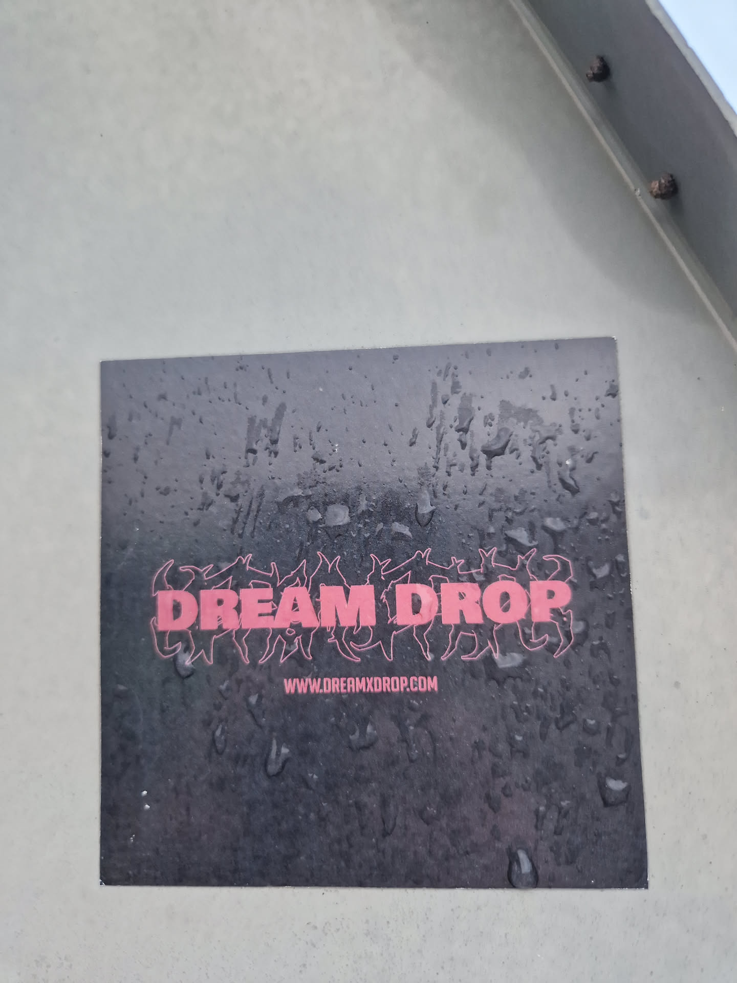 A black sticker with the words DREAM DROP in pink stylized text. Below is a website address: www.loremxdrop.com. The sticker appears to be wet, with water droplets visible on its surface.