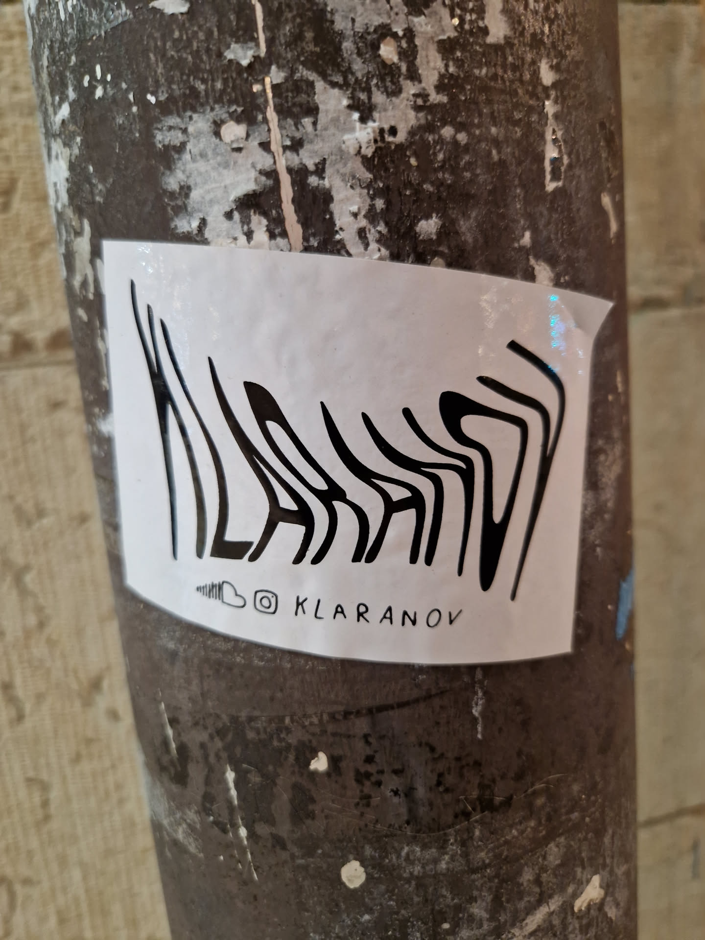 Black and white abstract design with the artist's Instagram handle at the bottom. The sticker is affixed to a weathered pillar.