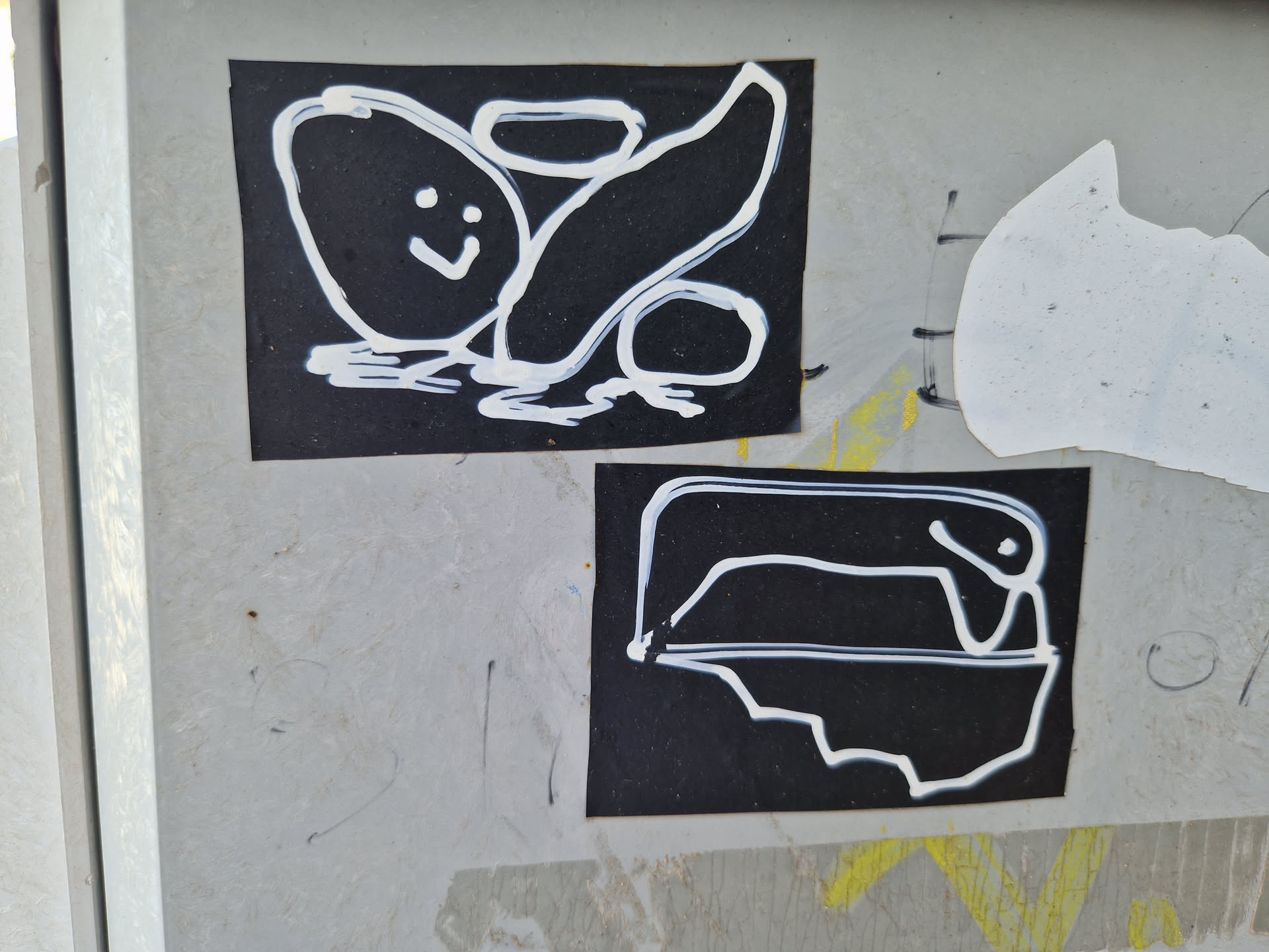 Two black stickers with simple white drawings. The top sticker shows a happy potato-like shape and a curved line. The bottom sticker depicts a simple line drawing of a creature or object in a container.  The style is childlike and playful.
