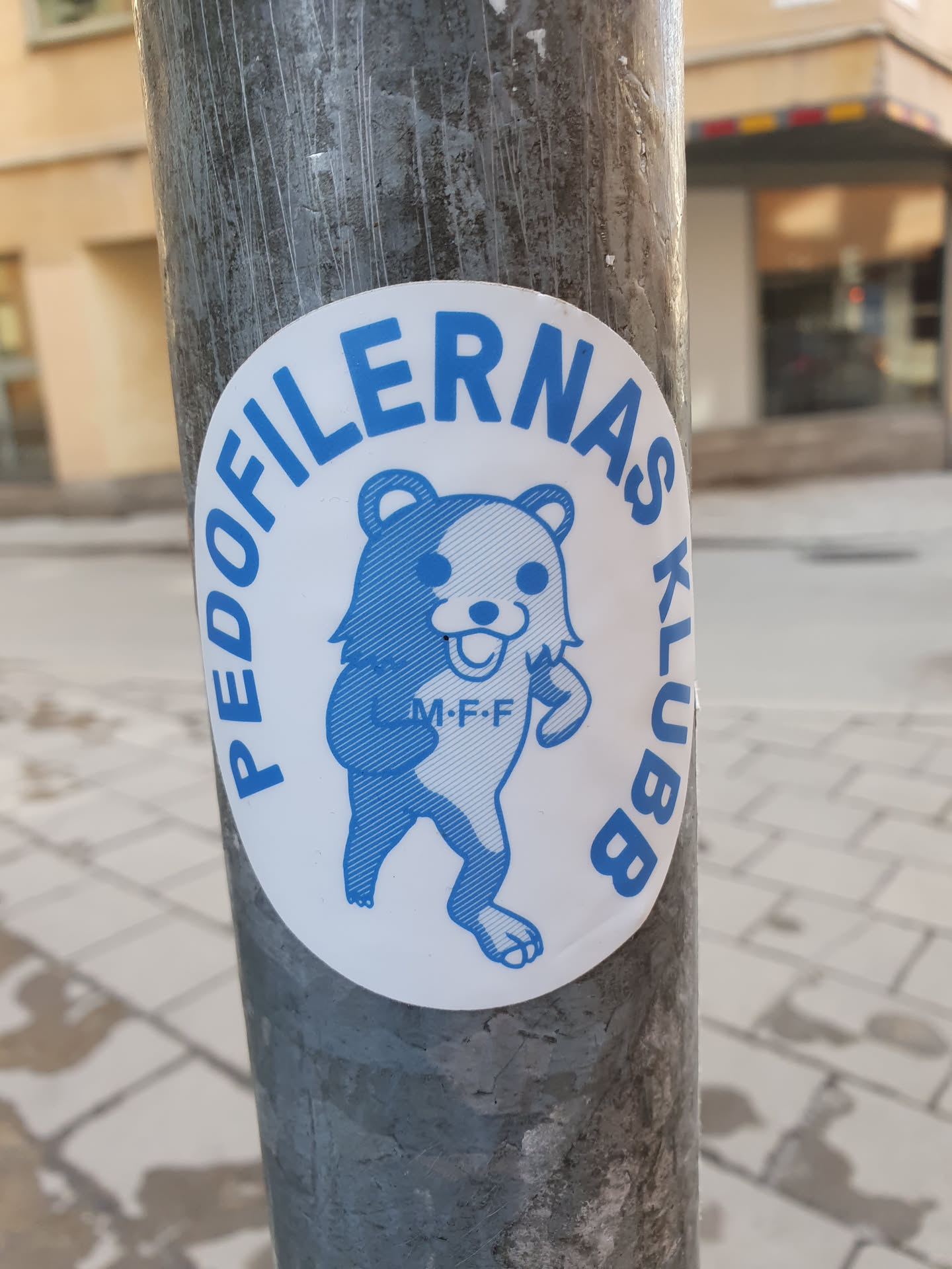 A round sticker affixed to a metal pole. The sticker features a cartoon bear wearing a shirt with the letters 'M-F-F'. It is surrounded by text that appears to read "Pedofilerna's Klubb." The text and imagery are in blue on a white background. The context suggests that the text might be Swedish, though the meaning is disturbing and potentially offensive.