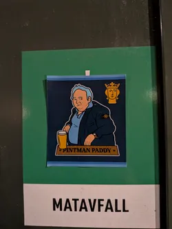 Street sticker A rectangular sticker on a dark navy background framed with light blue borders at the top and bottom. It features an illustration of 'Pintman Paddy' (Paddy Losty) with light blue-grey hair, seated at a bar with a pint of beer in front of him. He is wearing a dark jacket bearing the iconic Stone Island compass badge on the sleeve. In the upper right corner sits a golden silhouette emblem of Saint Eric, the symbol of the City of Stockholm. A gold banner at the bottom contains the text '★ PINTMAN PADDY ★'.