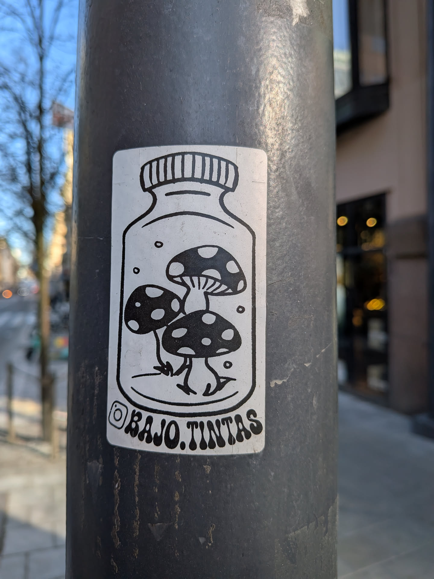 The sticker is a black and white illustration on a white background, shaped like a rectangle with rounded corners. It features a clear glass jar or bottle with a horizontally striped screw-top lid. Inside the jar, three stylized mushrooms are depicted with dark, spotted caps and lighter stems/gills. Small circular dots are scattered around the mushrooms within the jar. Below the jar, an Instagram logo is visible, followed by text.
