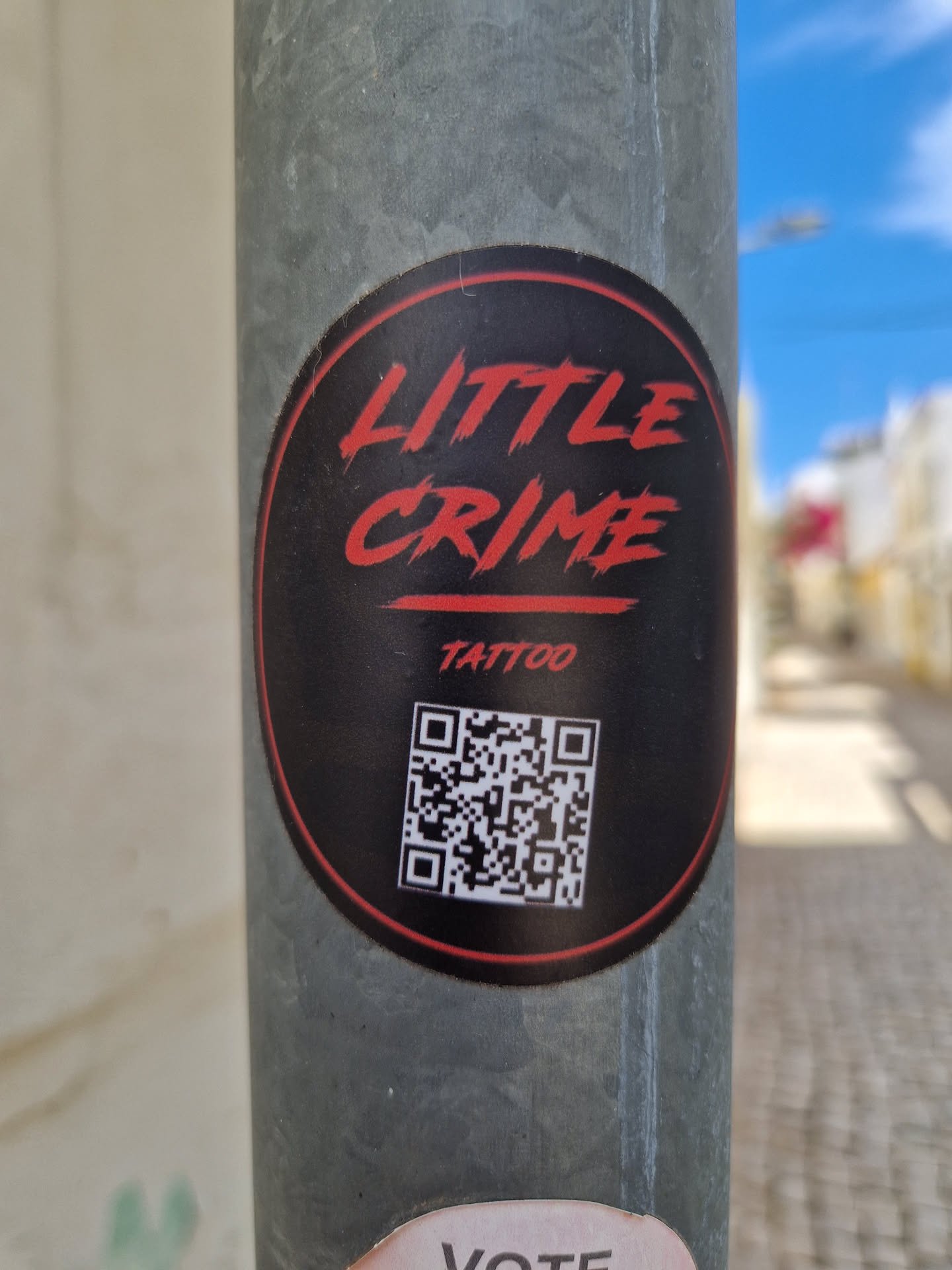 A round black and red sticker with the text "LITTLE CRIME TATTOO" and a QR code. The sticker is on a gray metal pole.