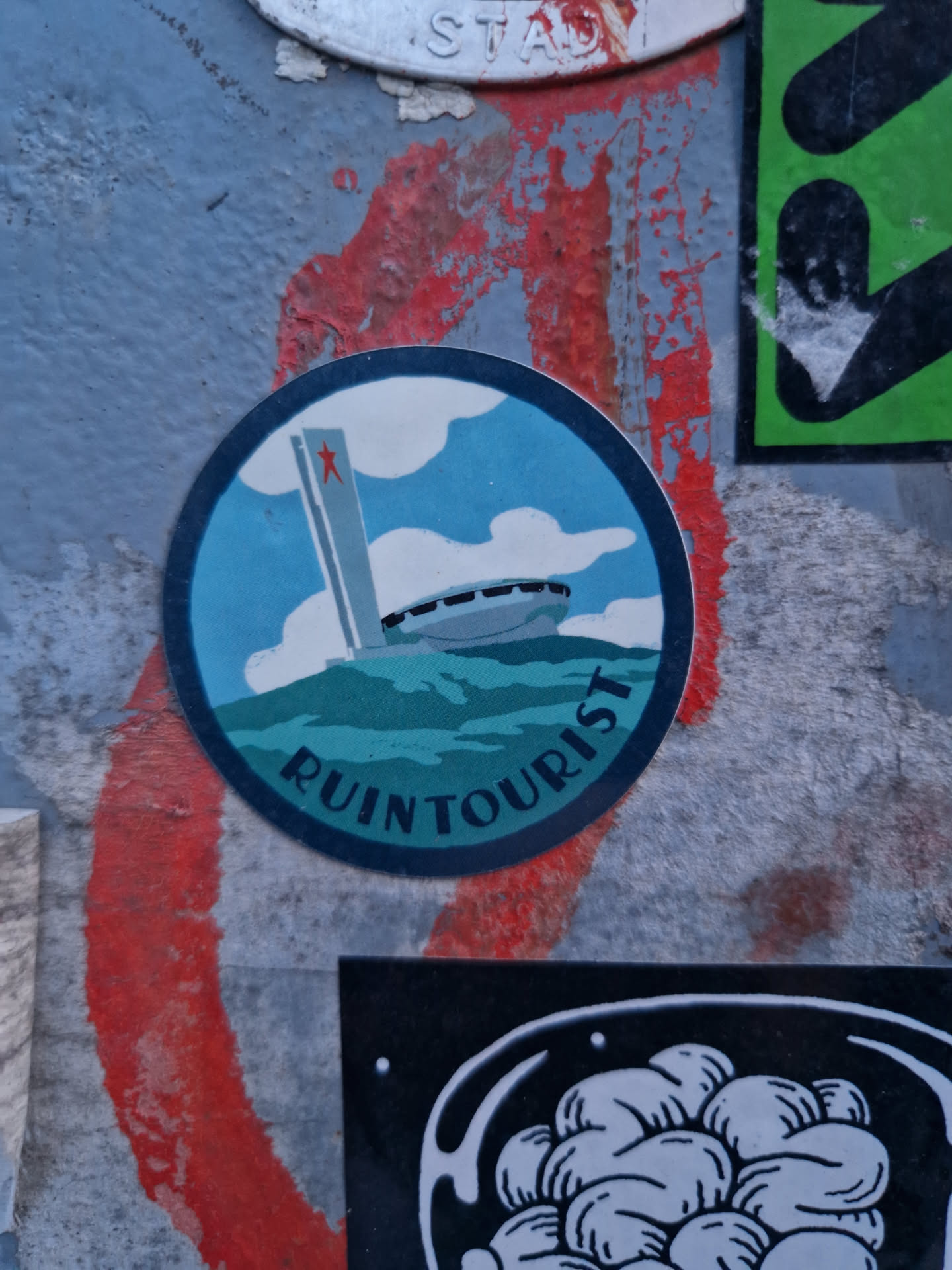 A round sticker featuring a stylized illustration of a building resembling the Palace of the Soviets, set against a teal background with a cloudy sky. The words "RUIN TOURIST" are written in a circular manner at the bottom. The sticker is adhered to a textured surface with other stickers around it.