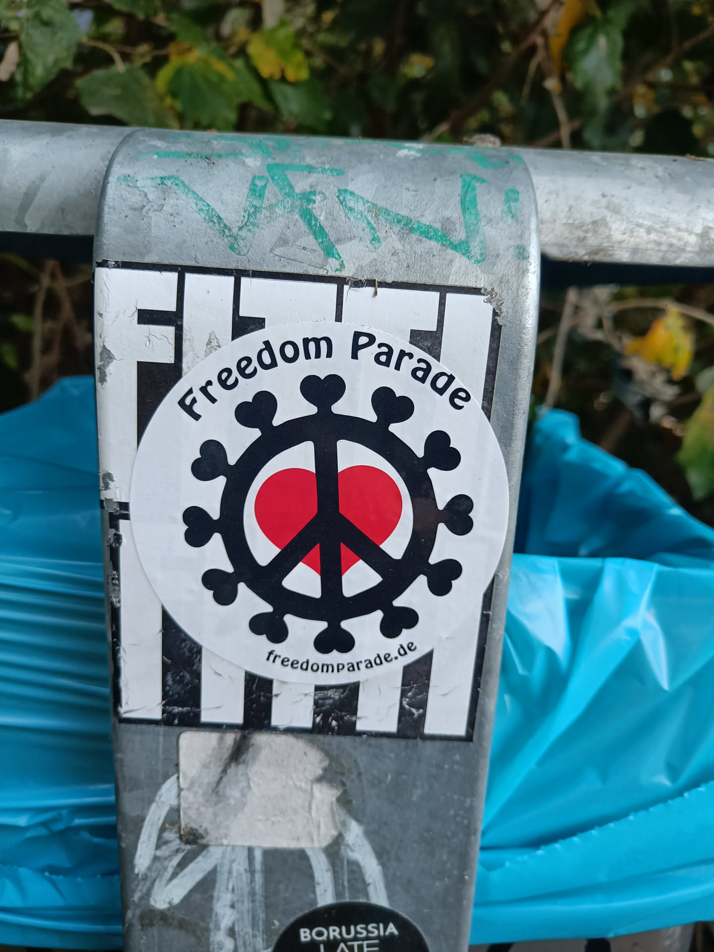 A round sticker with a black peace symbol encompassing a red heart, surrounded by a virus-like pattern.  The text "Freedom Parade" is printed above the design, and the website "freedomparade.de" is below. The sticker is affixed to a metal pole.