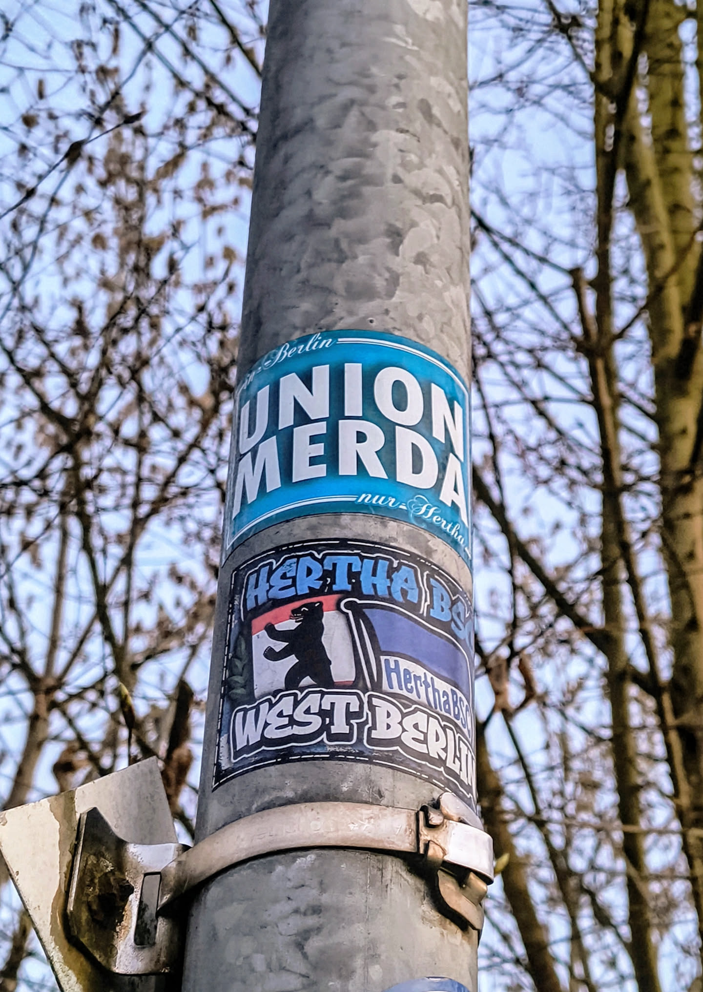 A metal pole with soccer related stickers attached to it. The stickers include text and graphics related to German soccer teams, including Hertha BSC West Berlin.