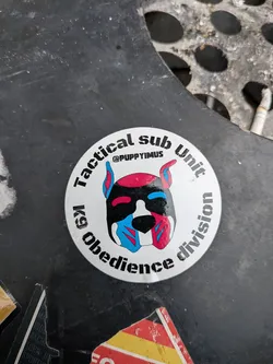 The sticker is circular with a white background. At the center is a stylized, cartoonish dog's face with bright red, black, and blue coloring. The dog has prominent ears, one pointing up and the other slightly bent. The design features geometric patterns and sharp lines. Text is arranged in a circular pattern around the dog's head. The top arc reads 'Tactical sub Unit' and the bottom arc reads 'k9 Obedience division'. Above the dog's face, centered, is the handle '@PUPPYIMUS'.