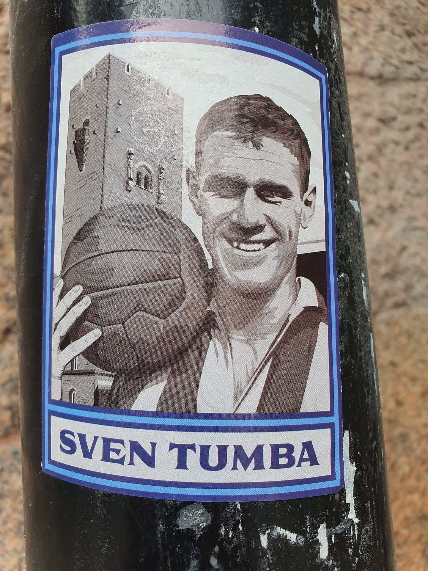 A grayscale sticker featuring Sven Tumba, a famous Swedish ice hockey player, holding a soccer ball with a building in the background.  The sticker has a blue border and "SVEN TUMBA" is written at the bottom.