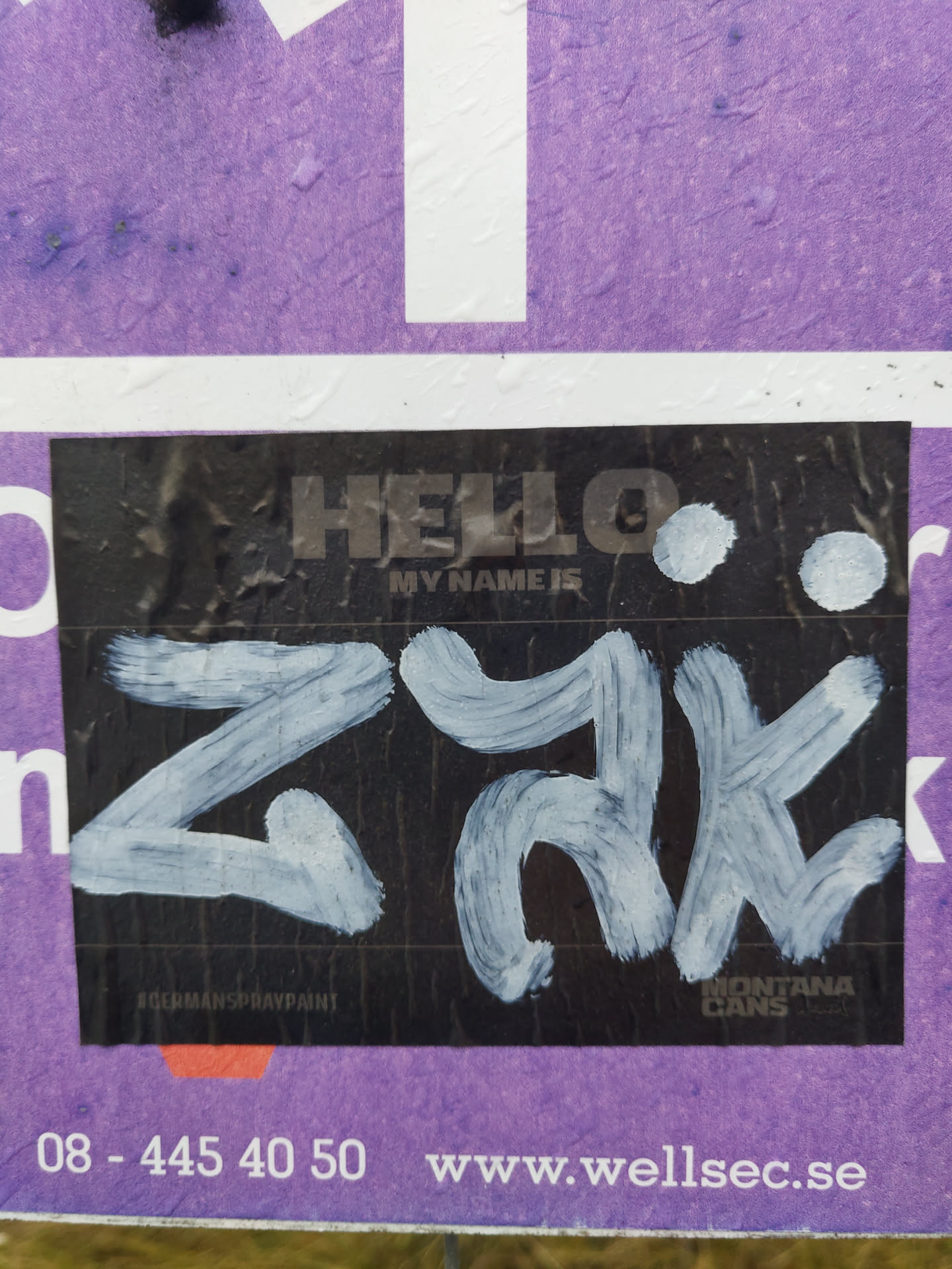 Black sticker with white paint. The text 'HELLO' and 'MY NAME IS' is printed on the sticker. Below is a stylized signature 'ZYKE' in white paint. The sticker also mentions 'ROCKMANS SPRAYPAINT' and 'MONTANA CANS'.