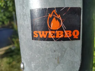 A sticker with an orange flame and the text "SWEBBQ" on a black background. The sticker is attached to a metal pole and shows some signs of wear.