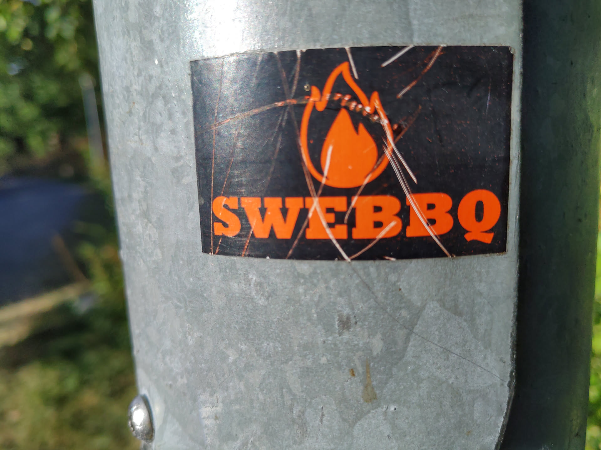 A sticker with an orange flame and the text "SWEBBQ" on a black background. The sticker is attached to a metal pole and shows some signs of wear.