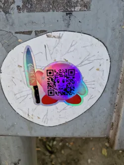 Holographic Kirby sticker with a QR code and a knife. The sticker is placed on a gray surface.