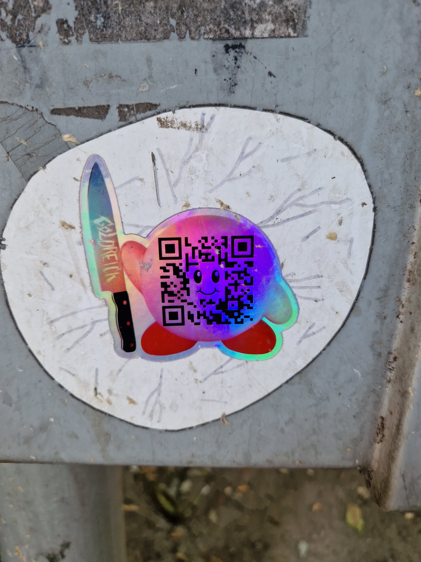 Holographic Kirby sticker with a QR code and a knife. The sticker is placed on a gray surface.