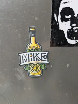 A sticker featuring an illustration of a bottle of lemon cello with the word MIKE in a stylized font. The bottom of the sticker says "Lemon Sunset."