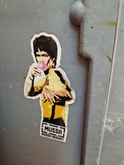 A sticker featuring a stylized image of Bruce Lee blowing a bubble of bubblegum.  The sticker includes the artist's name, Giorgio Mussati, and his brand, "Mussa".