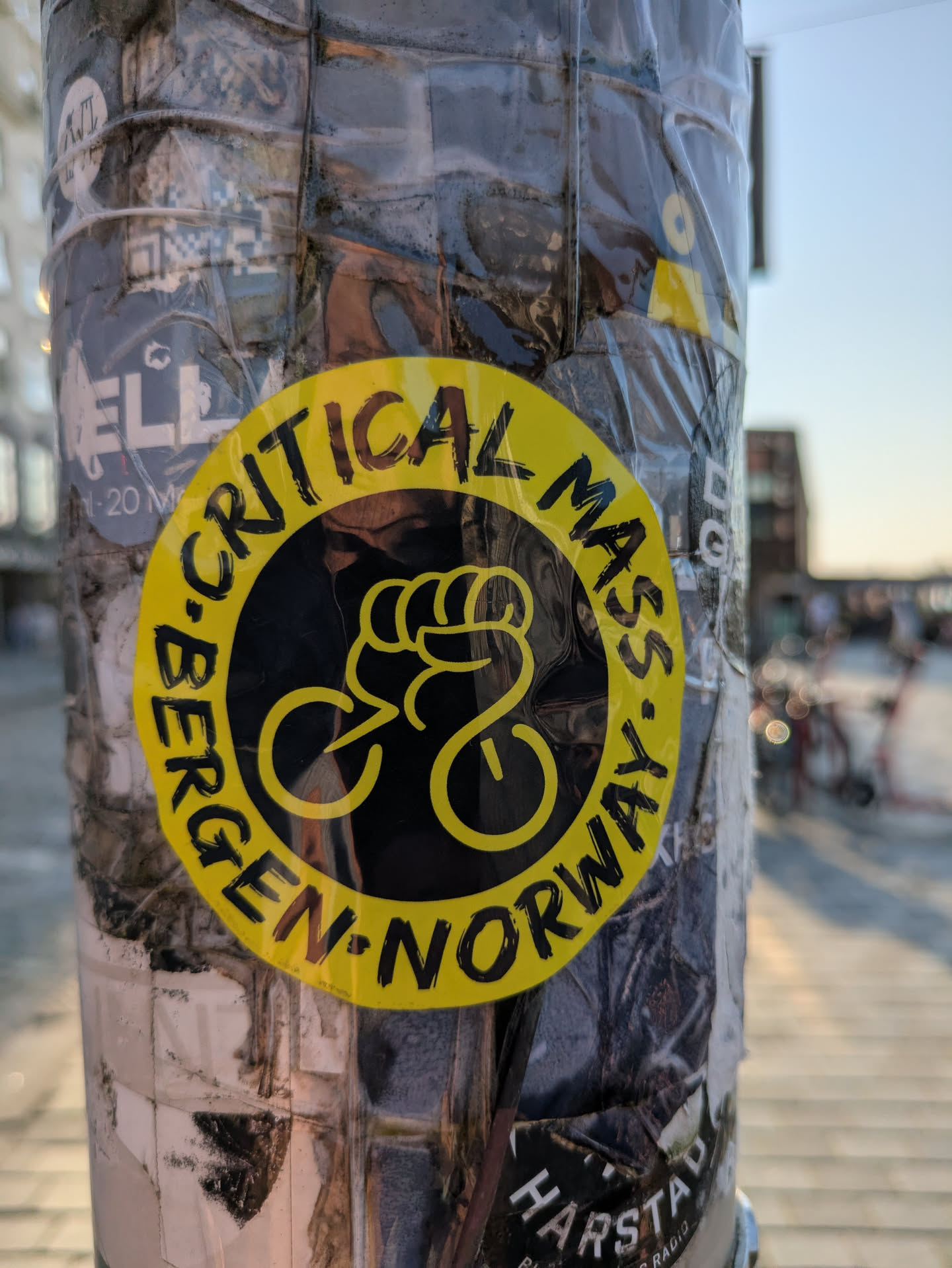 The prominent sticker is a circular design, primarily yellow with black text and an illustration. It features the words 'CRITICAL MASS' arching over the top and 'BERGEN NORWAY' arching along the bottom, both in a bold, brushstroke-like font. The center of the sticker displays a unique graphic: a yellow fist clenching what appears to be bicycle handlebars, with two circular shapes below representing wheels, forming a stylized bicycle silhouette. The sticker is affixed to a pole that is heavily covered with numerous other stickers, partially obscured by clear tape or plastic. The background shows an outdoor setting with blurred buildings and a street.