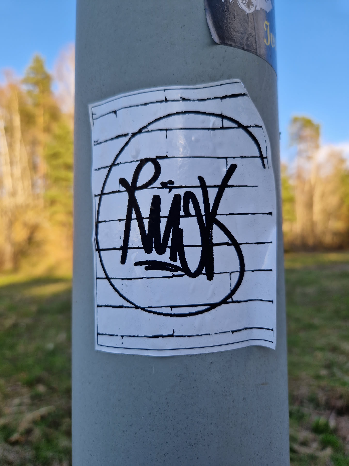 A sticker with graffiti-style artwork. The main element is a circular design with the word "RIÖK" written in black marker inside, set against a backdrop of brick pattern. The sticker appears to be affixed to a grey metal pole in an outdoor setting.