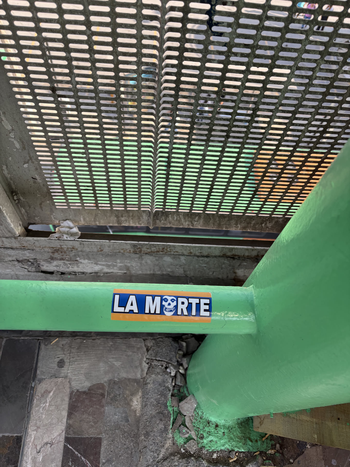 A rectangular sticker featuring the phrase 'LA MORTE' in bold white capital letters. The background is divided horizontally, with a dark blue section behind 'LA M' and a vibrant orange section behind 'ORTE'. The 'M' in 'MORTE' is cleverly integrated with a white skull design, reminiscent of the iconic Misfits band logo, complete with black eye sockets and nose. The sticker is applied to a bright green painted pipe.