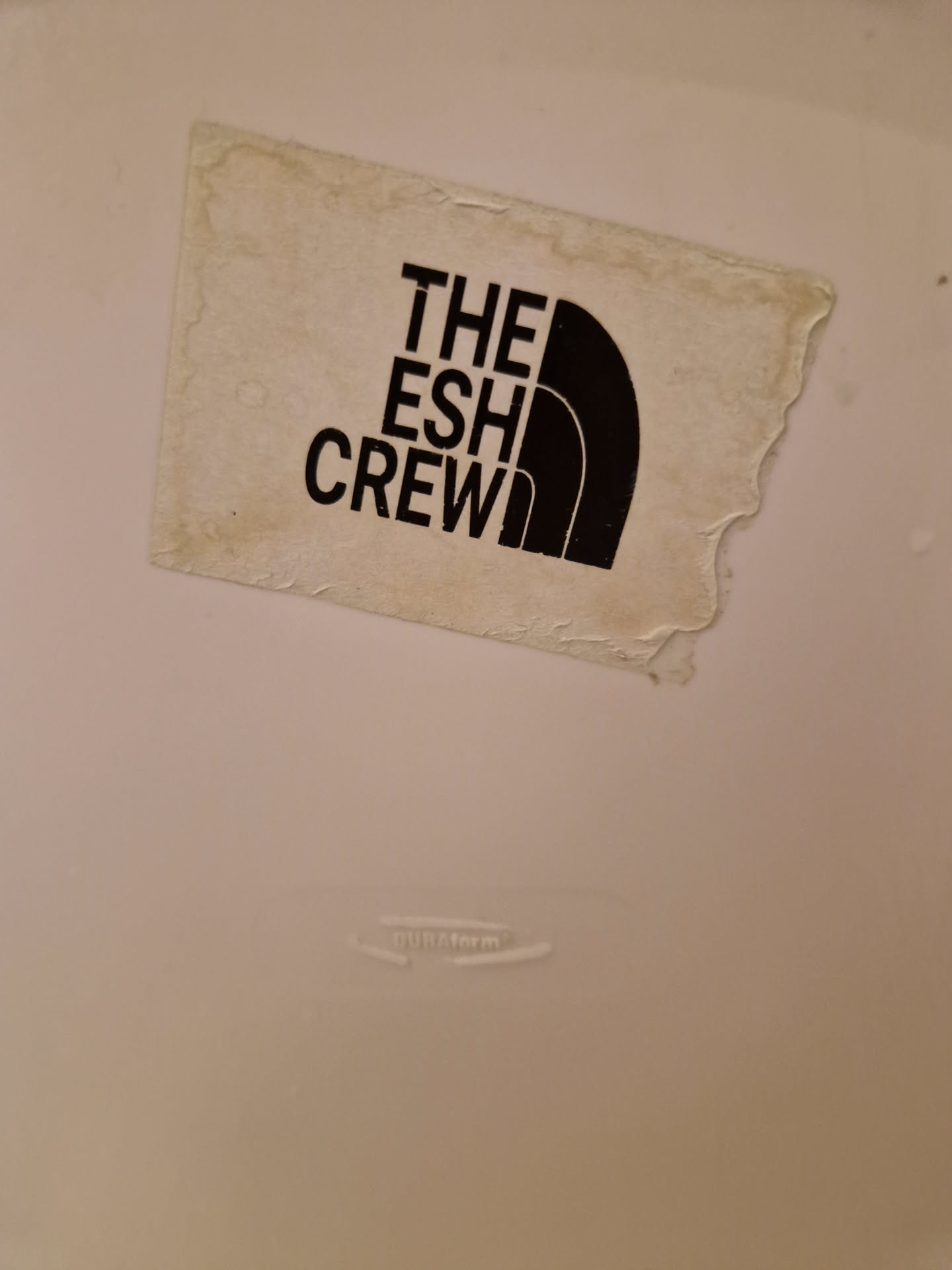 A worn-out, off-white sticker with black text and logo. The text reads "THE ESH CREW" and the logo is similar to the North Face logo.
