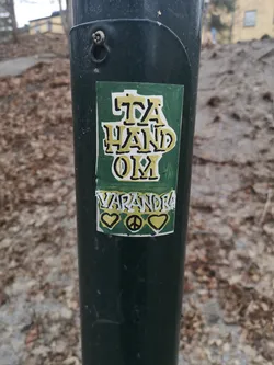 A rectangular, hand-drawn style sticker with a dark green background. The main text, 'TA HAND OM,' is prominently displayed in yellow with a black outline, followed by 'VARANDRA' in white with a black outline. Below the text, there are three symbols: a yellow heart, a black peace sign on a yellow circle, and another yellow heart, all outlined in black. The sticker has a somewhat rough, DIY aesthetic and is affixed to a dark green metal pole, showing minor signs of wear.