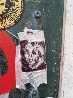 A dark maroon colored skull graphic on a beige sticker. The sticker is slightly torn and creased, adding to its worn look. The skull is depicted with an open mouth and seems to be wearing headphones or a similar object. The style of the drawing is detailed and slightly sketchy.