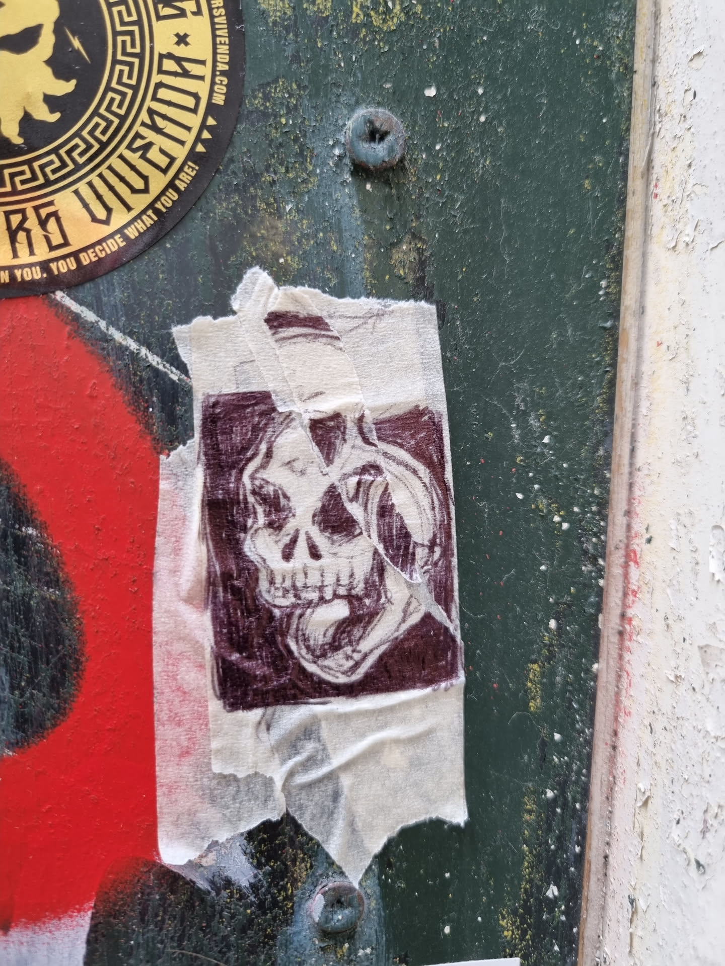 A dark maroon colored skull graphic on a beige sticker. The sticker is slightly torn and creased, adding to its worn look. The skull is depicted with an open mouth and seems to be wearing headphones or a similar object. The style of the drawing is detailed and slightly sketchy.
