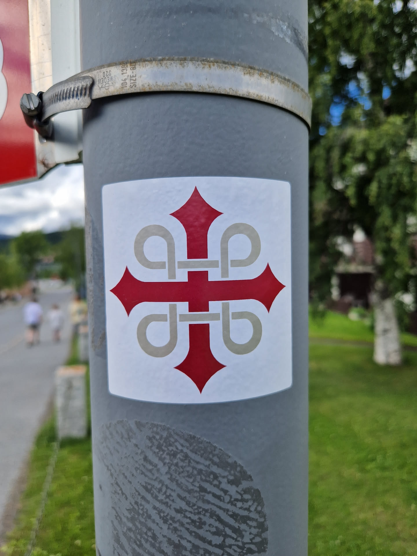 A square white sticker affixed to a grey pole.  The sticker features a stylized red cross with grey embellishments. The cross design is geometric and symmetrical. There is no visible text or QR code. The background is blurred, showing a street and some greenery.
