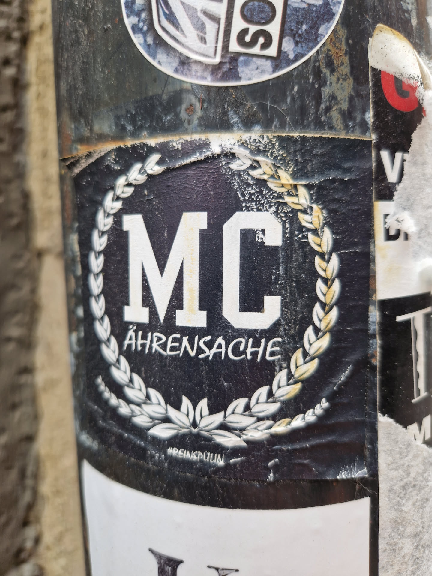 Black and white sticker with the letters MC in the center, surrounded by a laurel wreath. The text Ährensache is written below the letters. There is a small text at the bottom that seems to be a signature or a watermark.