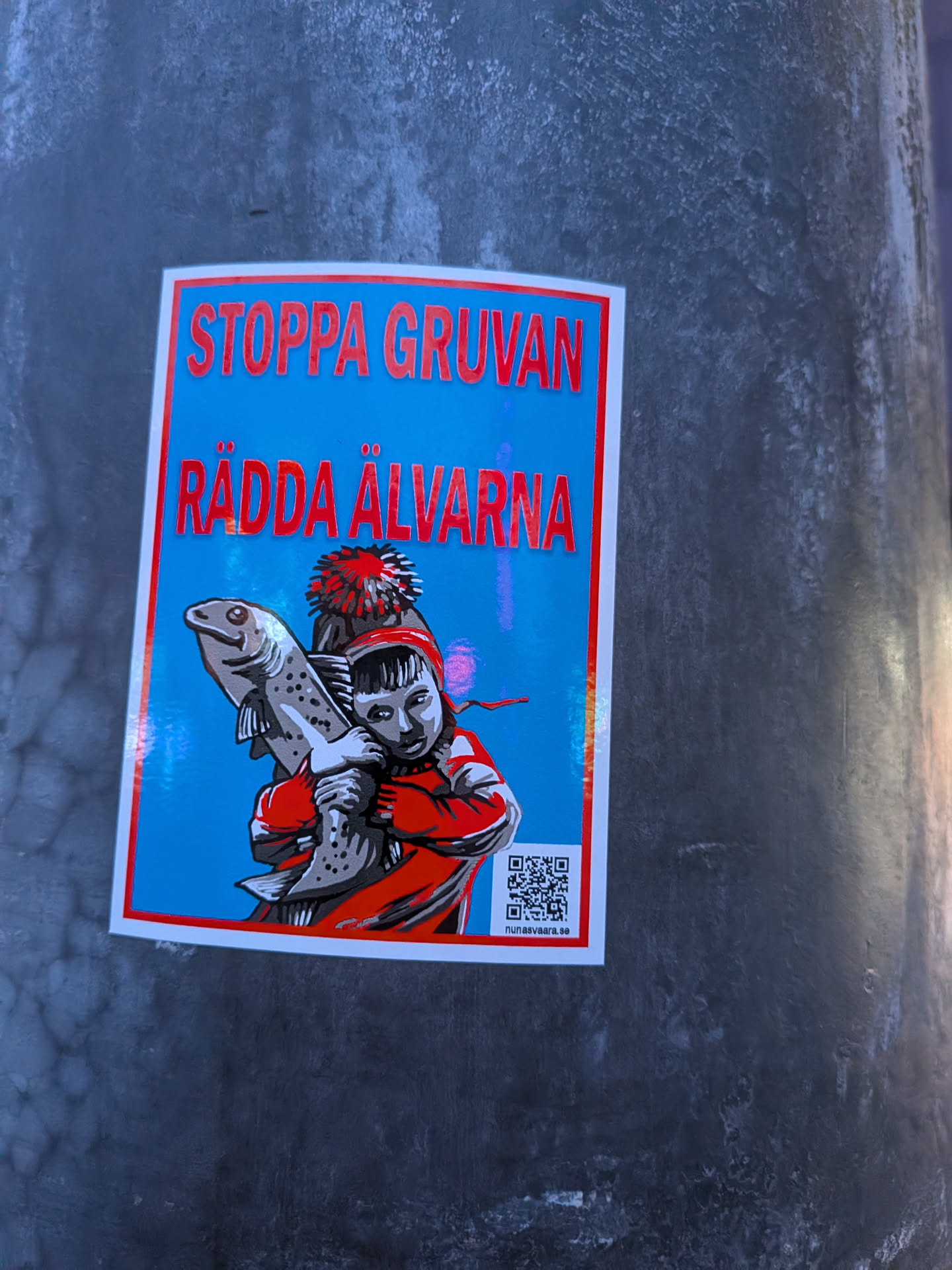 The sticker features a vibrant blue background with a red border. In the center, a stylized illustration depicts a child wearing a red jacket and a red and white pom-pom hat, holding a large, speckled fish. The child has dark hair and a determined expression. The text 'STOPPA GRUVAN' and 'RÄDDA ÄLVARNA' is prominently displayed in bold red lettering at the top of the sticker. Below the illustration, a QR code is visible, and beneath that, the text 'nunäsvaara.se'. The overall style is graphic and impactful, conveying a message of environmental concern.