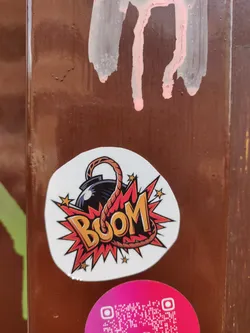 A round sticker featuring a cartoon bomb with a lit fuse and the word "BOOM" in a stylized font. The design is vibrant and colorful, with a red and orange color scheme. The sticker is affixed to a brown wooden surface.