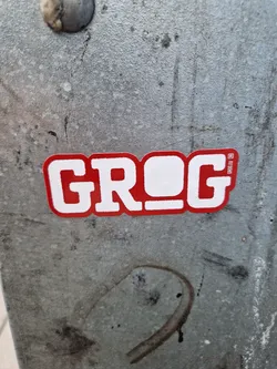Red and white sticker with the word Grog on it. The sticker has a slightly worn appearance and is placed on a grey metallic surface.
