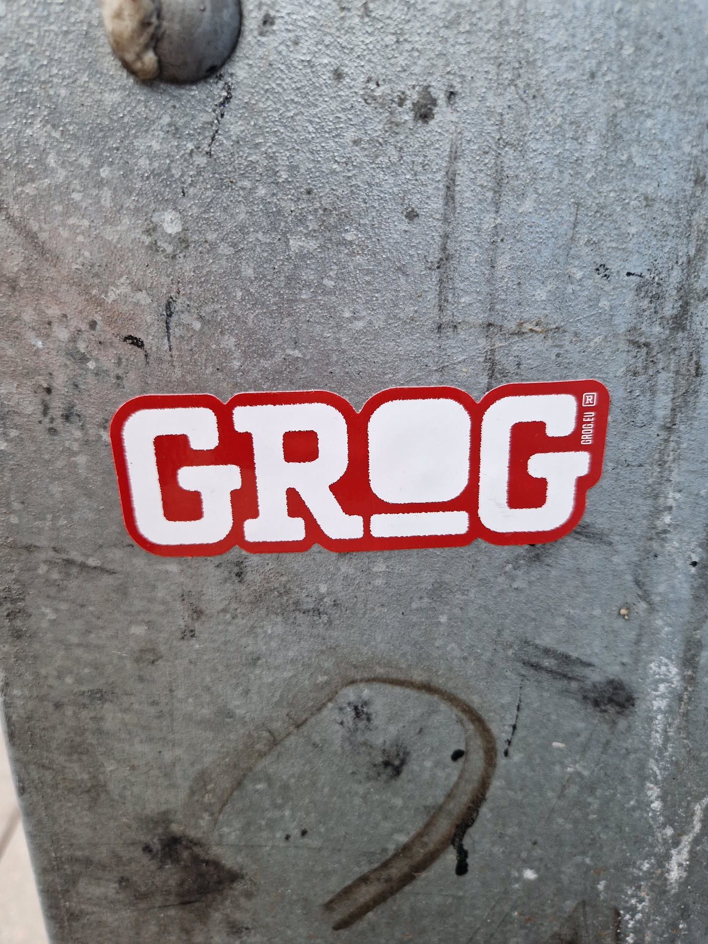 Red and white sticker with the word Grog on it. The sticker has a slightly worn appearance and is placed on a grey metallic surface.