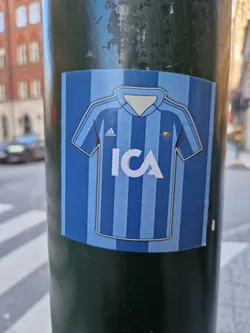 A sticker featuring a stylized illustration of a light blue football jersey with vertical stripes. The jersey has the Adidas logo and the ICA logo in the center. The sticker is affixed to a dark green pole.
