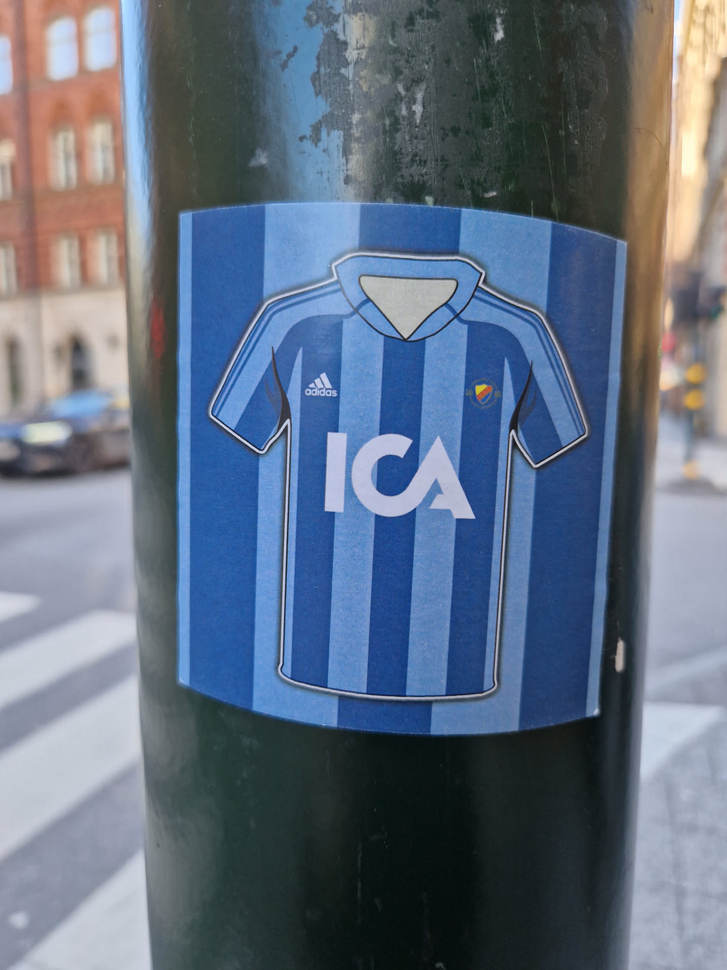 A sticker featuring a stylized illustration of a light blue football jersey with vertical stripes. The jersey has the Adidas logo and the ICA logo in the center. The sticker is affixed to a dark green pole.