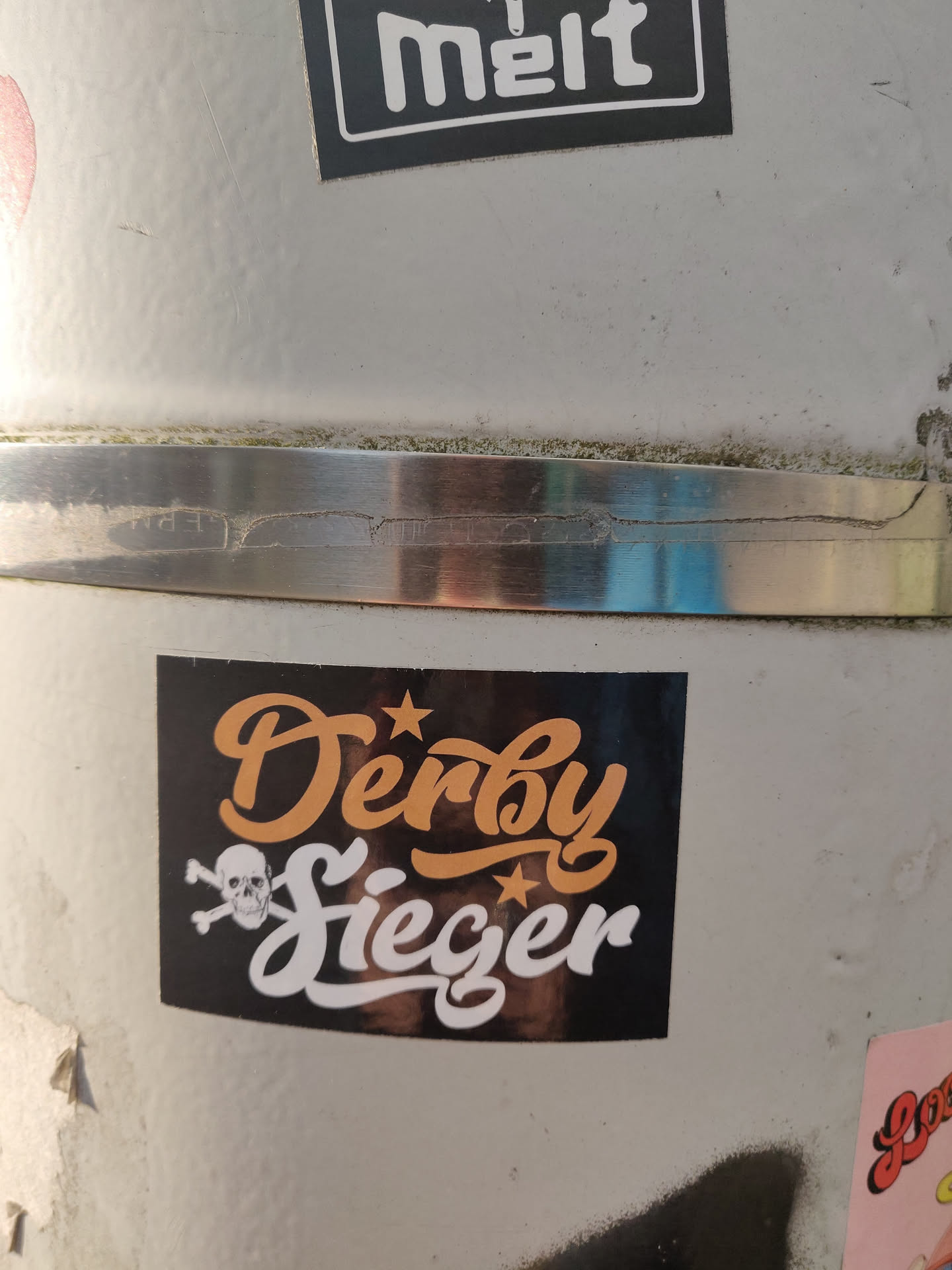 A black sticker with the text "Derby Sieger" in orange and white stylized lettering.  A skull and crossbones is featured below the text. The sticker appears to be affixed to a metal surface.