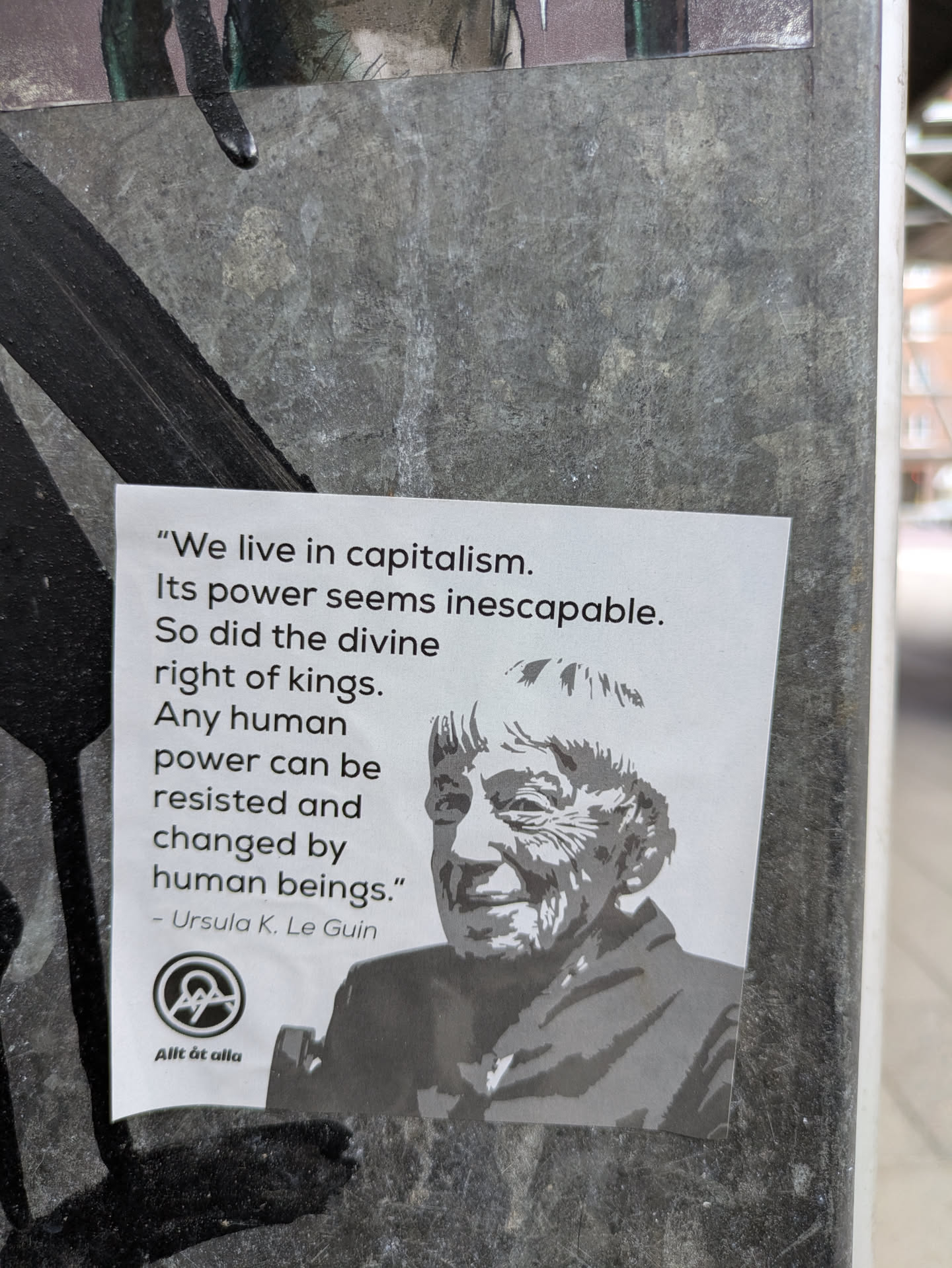 The sticker is a black and white design featuring a quote from Ursula K. Le Guin about capitalism and resisting power. It also shows a portrait of the author and includes a logo and the text 'Allt åt alla'. The sticker is affixed to a textured, possibly concrete or stone, surface.