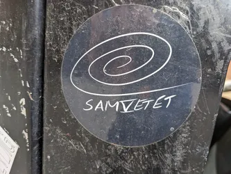 A round, dark-colored sticker features a hand-drawn spiral design in white. Below the spiral, the word 'SAMVETET' is written in a stylized, handwritten font. The sticker is affixed to a dark, textured surface, possibly metal.