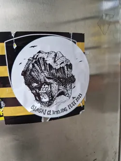 Black and white illustration of a cliff with a house at its base. The sticker is circular with a black border and yellow and black stripes on the background.