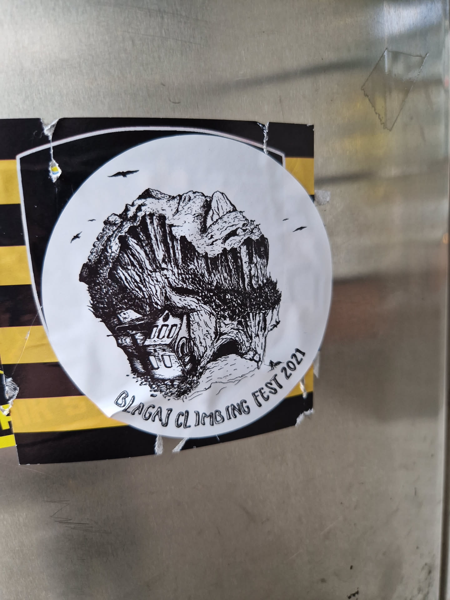 Black and white illustration of a cliff with a house at its base. The sticker is circular with a black border and yellow and black stripes on the background.