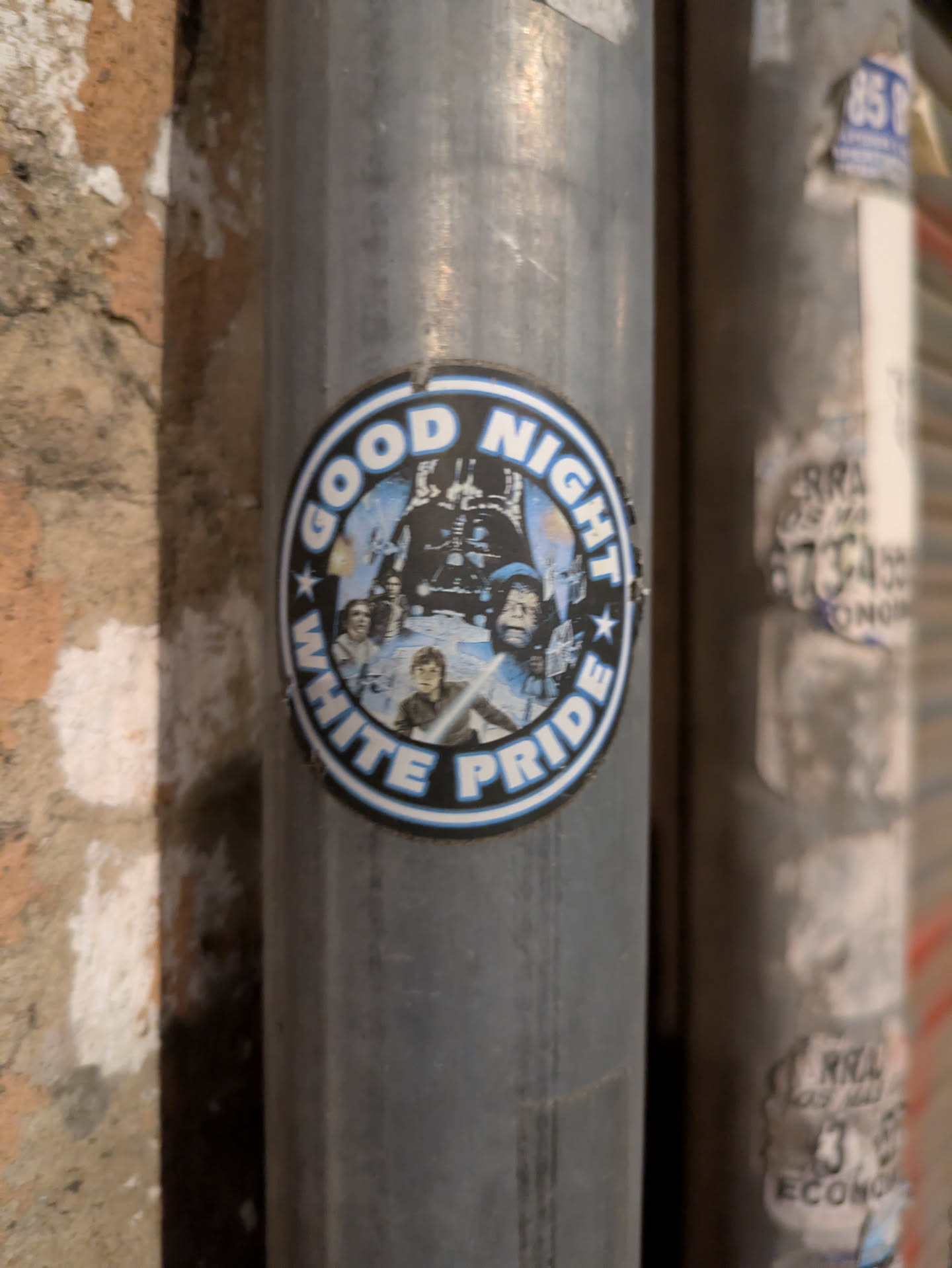 A round sticker stuck on a metal pole. The sticker shows characters from Star Wars movie and the words 'GOOD NIGHT WHITE PRIDE' in a circular layout. The words are written in white with a blue outline.