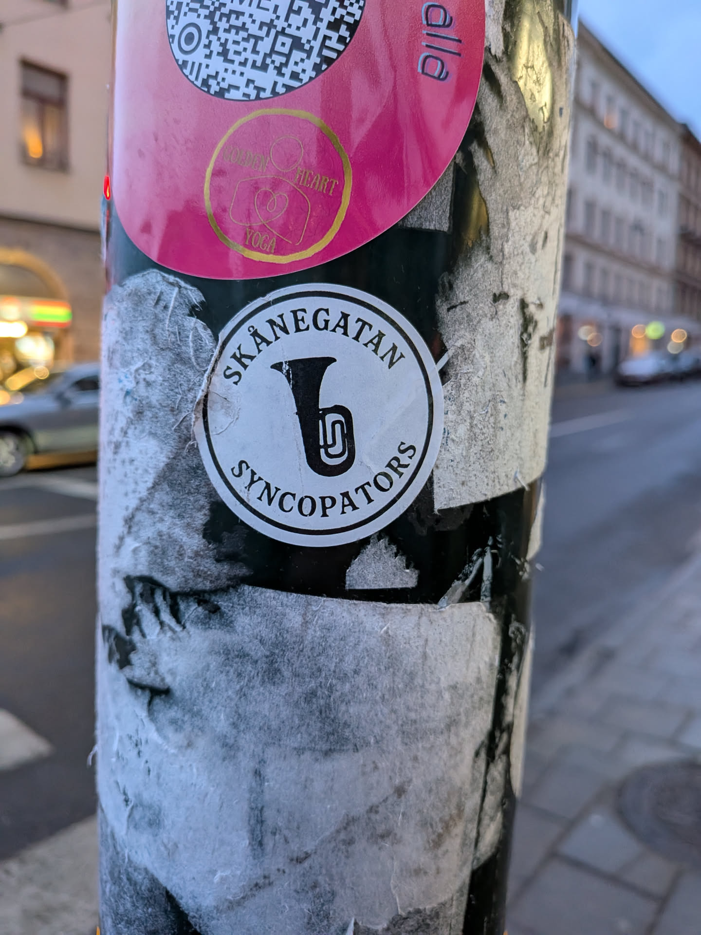 The sticker is circular with a white background and a thin black outer border. At its center is a stylized black illustration of a tuba or a similar brass instrument, depicted with a simple, clean line art style. Arcing above the instrument, following the curve of the circle, is the word "SKÅNEGATAN" in black sans-serif capital letters. Below the instrument, also arcing along the circle's bottom edge, is the word "SYNCOPATORS" in similar black sans-serif capital letters. The overall design is monochrome, clean, and has a classic band logo aesthetic.