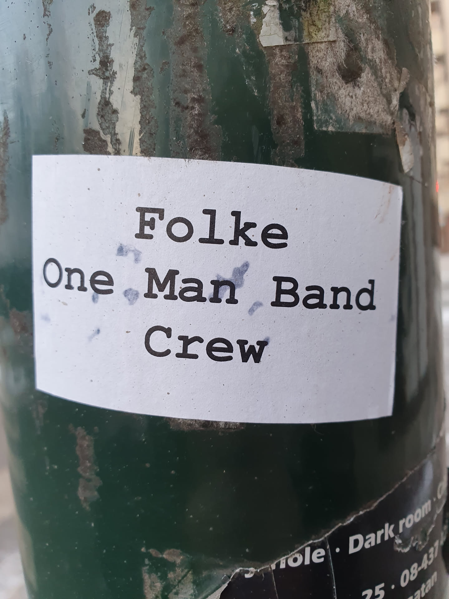 A sticker featuring the text "Folke One Man Band Crew" in a typewriter-style font. The sticker is affixed to a dark-green pole. 