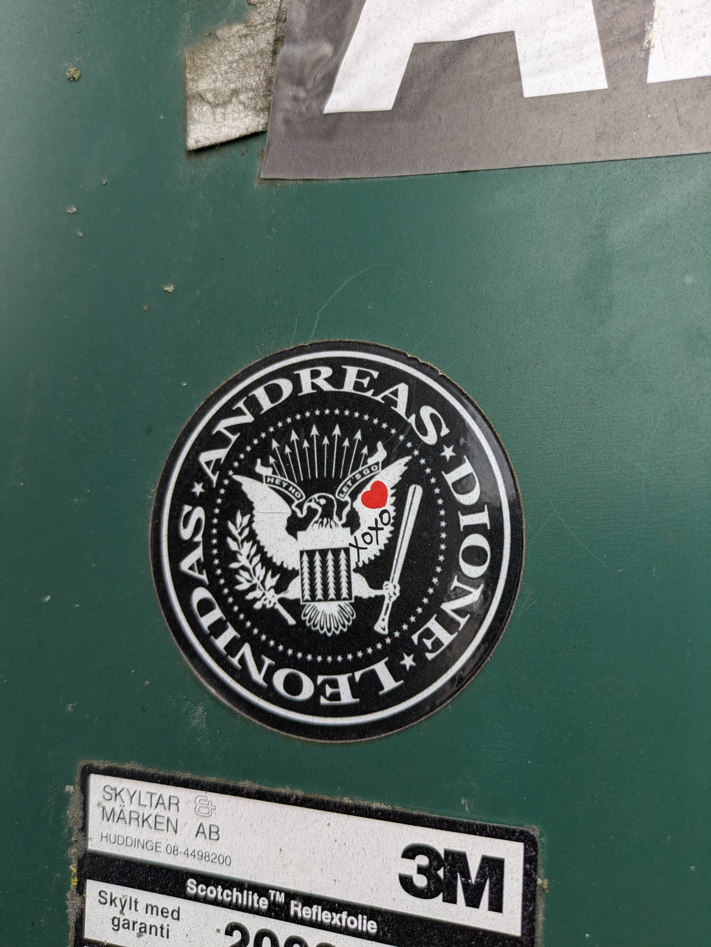 A circular black and white sticker affixed to a dark green surface. The central design is an eagle, reminiscent of a presidential seal, holding arrows in one talon and a branch in the other. Above the eagle's wings are the phrases 'HEY HO' on the left and 'LET'S GO!' on the right, a direct reference to The Ramones' song. A small red heart and 'xoxo' are visible near the arrows. The outer ring of the sticker displays the text 'ANDREAS DIONE' at the top and 'LEONIDAS' at the bottom, separated by stars. The sticker combines formal heraldry with an irreverent punk rock aesthetic.