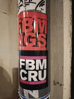 Two stickers on a pole, one says FBM HGS in red and black, and the other says FBM CRU in white, pink and black. 