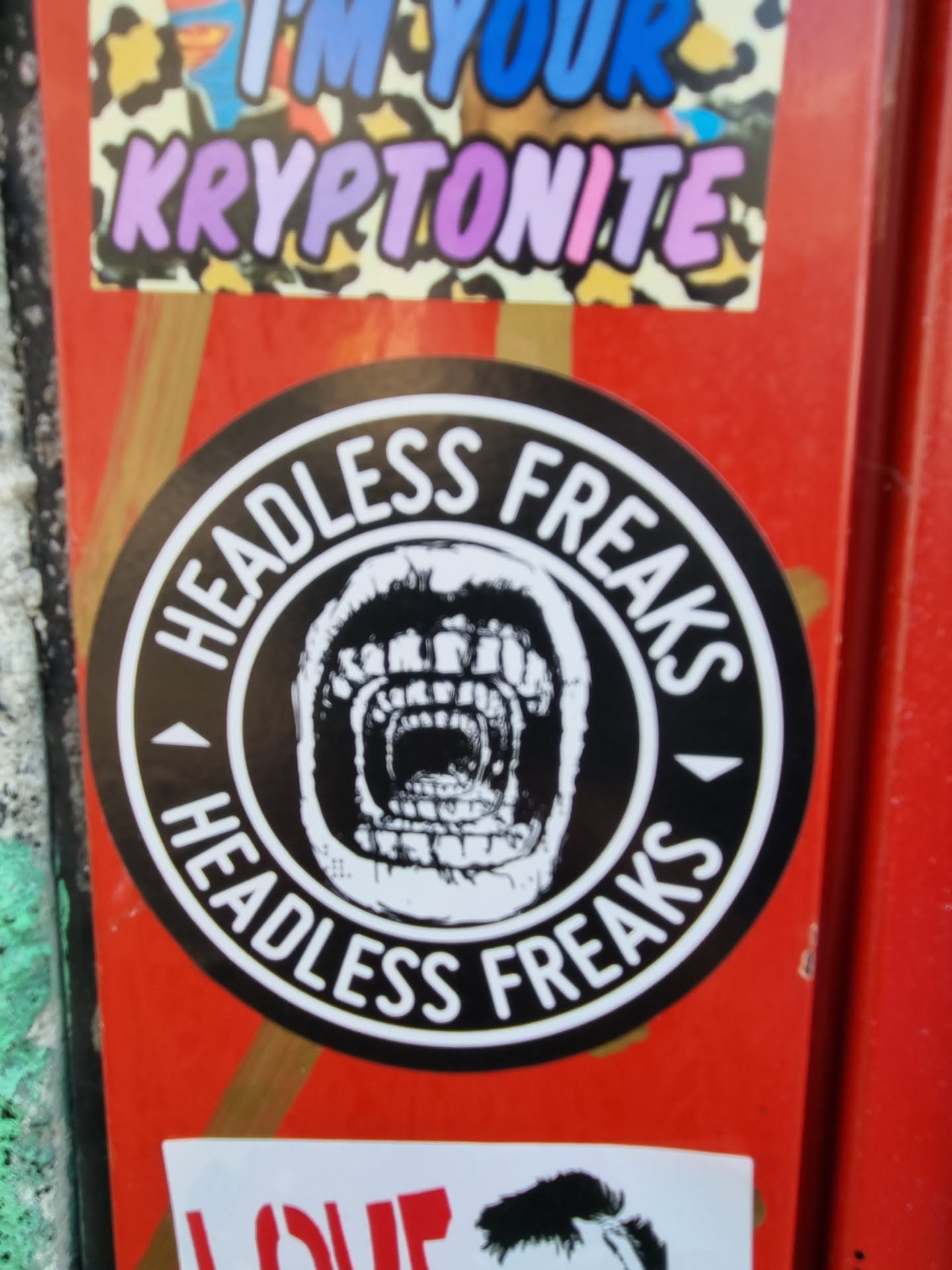 A black and white round sticker with the words "Headless Freaks" in a circular design.  The center features a graphic of a screaming skull with multiple sets of teeth.  The sticker is affixed to an orange surface with other stickers around it.