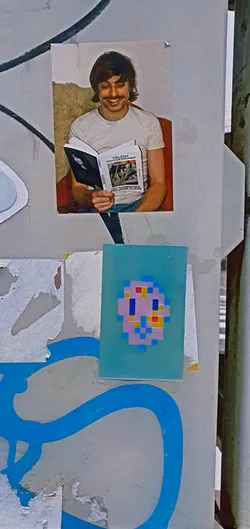 A turquoise sticker featuring a pixelated face, made up of pink, blue, yellow, and red squares, is adhered to a weathered wall.