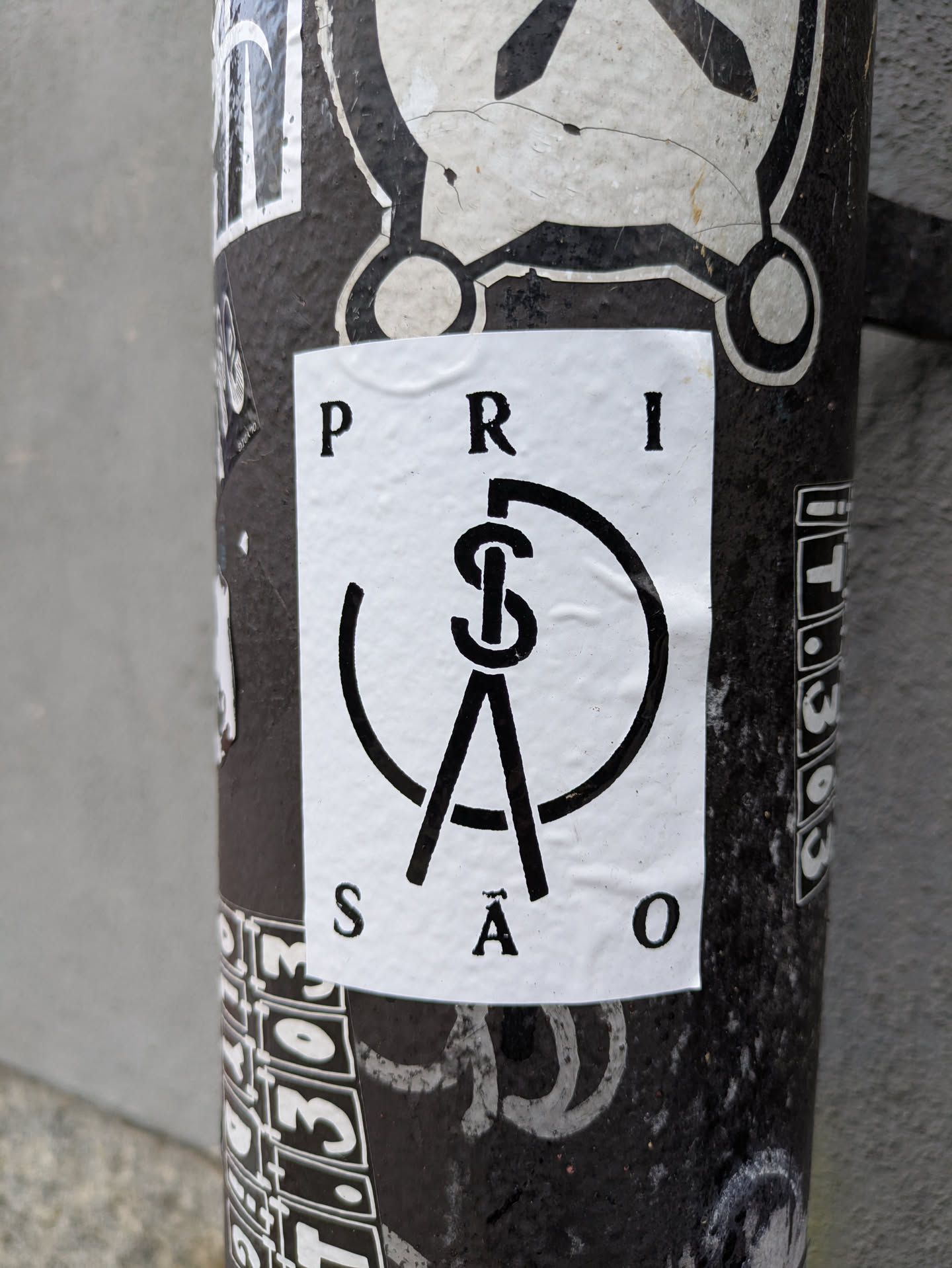 Black and white sticker with the word PRISÃO (Prison in Portuguese) forming a circle around a symbol that looks like a compass or a pair of dividers. The style is minimalist and geometric.
