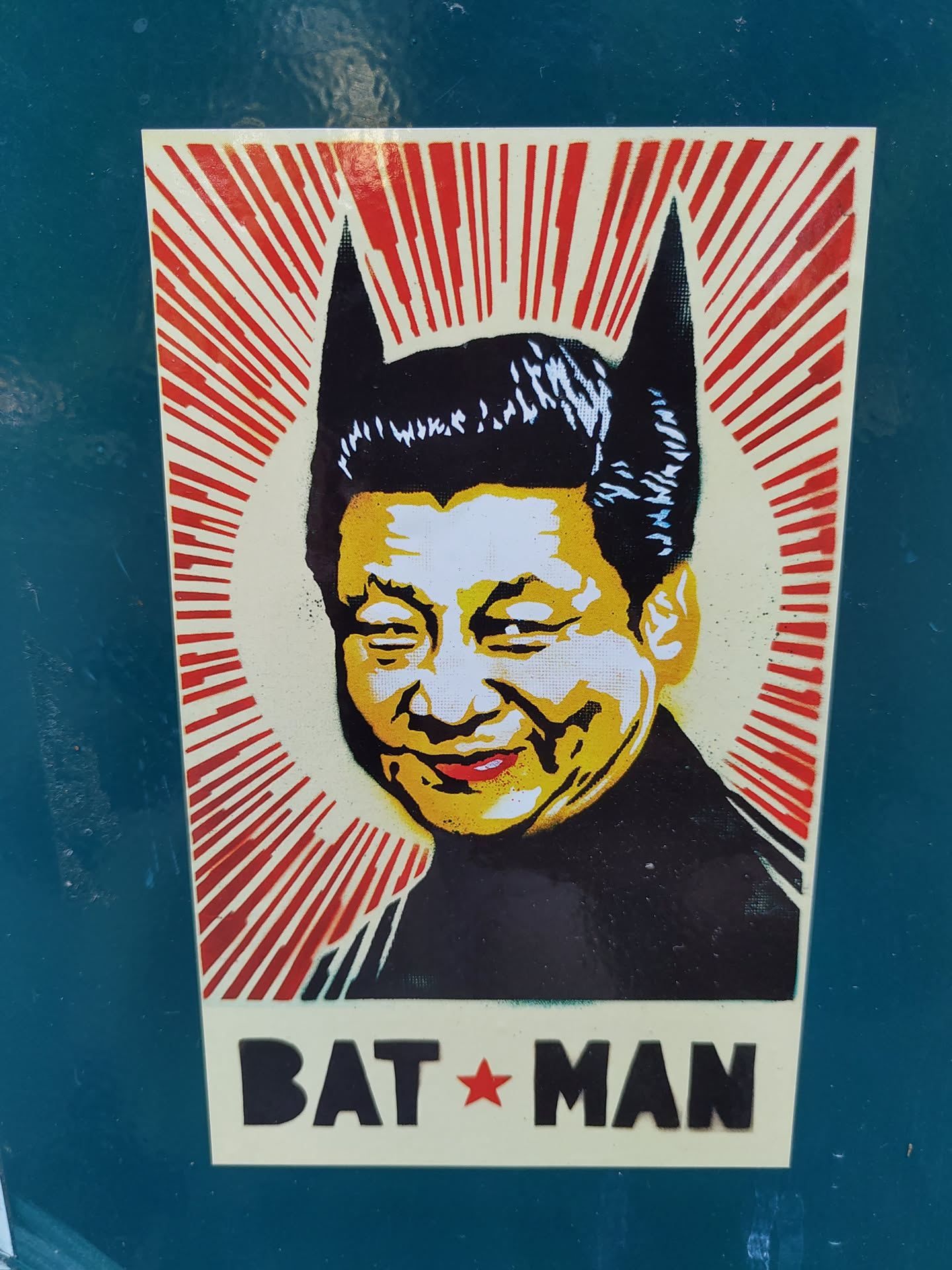 A sticker depicting Xi Jinping, the Chinese president, with Batman's ears. The style is reminiscent of propaganda posters, with a red and white sunburst background.  The sticker has the text 'BAT*MAN' at the bottom.
