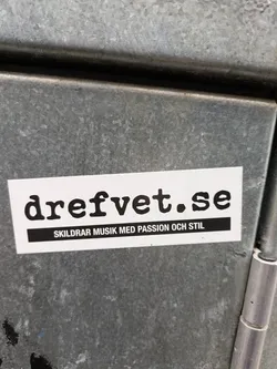A white sticker with black text on a gray metal surface. The text on the sticker says "dreftvet.se" in a large font and "Skildrar musik med passion och stil" in a smaller font below.